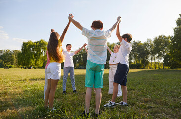 Fototapeta premium Outdoor children's games. Children standing in circle holding hands and playing games together having fun in park. Happy preteen boys and teenage girls in summer casual clothes raise hands together.