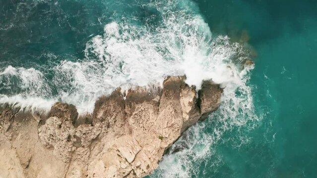 Viewed from an overhead drone, the scene unveils striking turquoise waters, with white waves crashing against a vibrant brown rock along Gozo's coast, sending sea spray soaring. LuPa Creative.