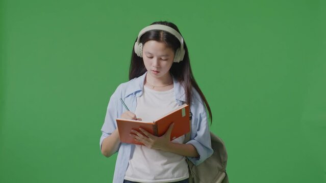 Asian Teen Girl Student With A Backpack Wearing Headphones And Taking Note On Notebook While Standing In The Green Screen Background Studio
