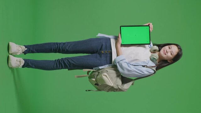 Full Body Of Asian Teen Girl Student With A Backpack Smiling And Showing Green Screen Tablet To Camera While Standing In The Green Screen Background Studio
