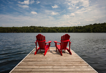Two red adirondack chairs on dock overlooking lake on sunny day.
