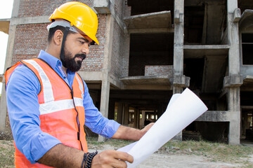 Young Indian male civil engineer or architect wearing helmet and vest holding paperwork blueprint at construction site.