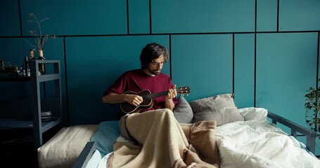 A brunette man in a dark red shirt lies on a sofa wrapped in a blanket and plays the guitar against a turquoise wall