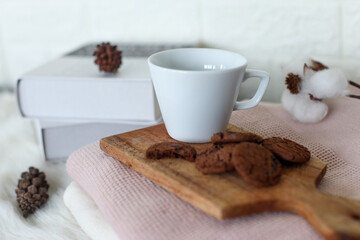A white cup of coffee or hot chocolate and chocolate chips cookies on wooden tray with pine cones and books on the background