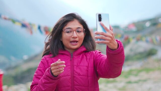 Portrait of excited young Indian woman talking on video call with vivid expressions. Girl connects with family with smart phone. Tourist making vlog in mountains.
