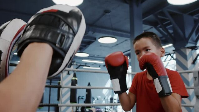 Portrait of a asian kid boy in boxing gloves is working out on a series of punches to the boxing paws of his trainer close up. Child sports training in an indoor gym