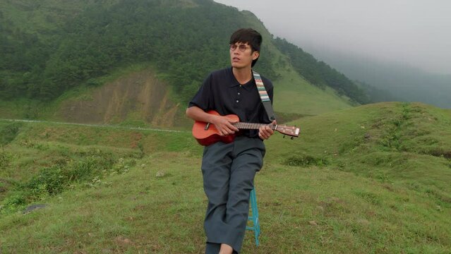 Young cool male playing the Hawaiian ukelele on tropical hilltop. Aerial dolly out