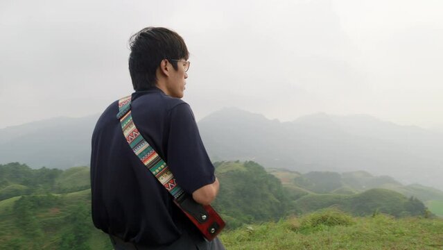 Melodramatic asian male playing the ukelele on tropical hilltop of Southeast Asia. Aerial