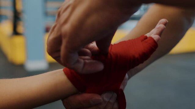 Coach wraps kid boxer hands with protection band. Trainer help boy to put on hand protective bandage before fight. Closeup of coach helping cover unrecognisable male hands with band