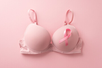 Uniting for breast cancer awareness. Top view photo showcasing a bra with attached pink ribbon symbol on soft pink backdrop. Perfect for promotional purposes or advertisements - obrazy, fototapety, plakaty