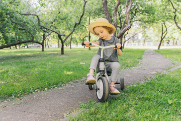 A little girl rides along a path in an apple orchard. The girl is wearing a straw hat. The weather is fine outside, the sun is shining. The frame is dynamic. The baby turns the steering wheel.
