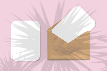 An envelope with two sheets of textured white paper on pink background. Mock up with an overlay of palm tree shadows. Natural light casts shadows from the leaves of a tree branch. Horizontal