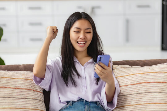 Excited happy young Asian woman holding smart phone device, looking at mobile smartphone screen gesturing yes, she received good news sitting on sofa at home