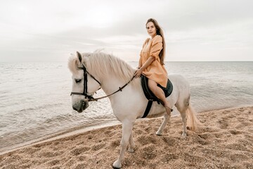 A white horse and a woman in a dress stand on a beach, with the sky and sea creating a picturesque...