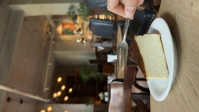Eating delicious basque burnt cheesecake in a cafe. Taking a piece of cake with a dessert fork. Atmosphere of a coffee shop in bokeh. Lifestyle vertical video.