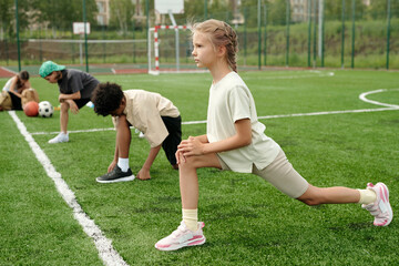 Side view of cute blond schoolgirl in activewear doing stretching exercise for legs during warm up before physical training lesson on stadium
