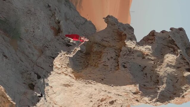 A drone flies around a young woman in a red dress standing on a cliff in the middle of the desert sands