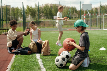 Side view of schoolboy in activewear choosing ball for playing sports game while standing on his knees on green lawn of football field