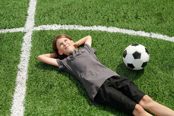 Above angle of cute restful schoolboy in activewear relaxing on green lawn of football field at...