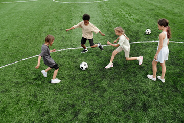 Group of intercultural schoolchildren following soccer ball while running along green football field during game or lesson of physical training