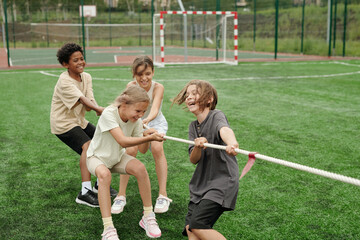 Cheerful schoolkids in activewear laughing while pulling rope during sports competition with...