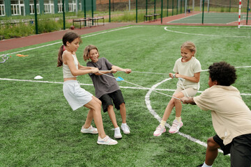 Obraz premium Four cute intercultural schoolkids in activewear pulling rope at stadium while standing on green football field and training before game