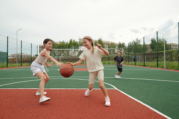 Two cute blond schoolgirls in activewear dribbling ball while running along field or stadium towards basket on background of their classmate