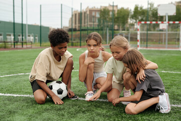 Four schoolchildren in activewear standing on knees on green football field or sports ground and having break between outdoor trainings