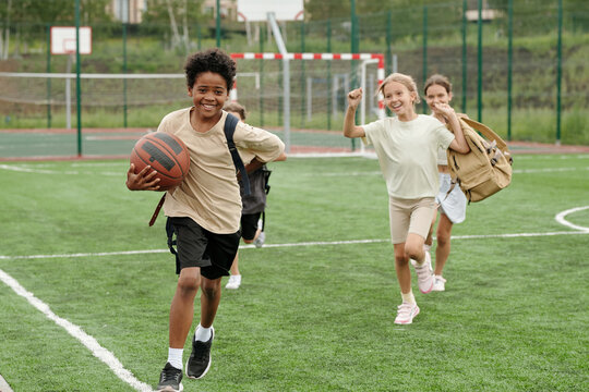 Two cute blond girld running after African American schoolboy with ball while having fun at stadium and enjoying pastime after school - Powered by Adobe