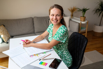 young adult student brunette woman wearing summer dress sits at the desk at home doing a homework and smiling.