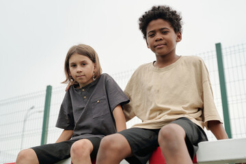 Two cute intercultural schoolboys in t-shirts looking at camera while sitting on top of goal net...
