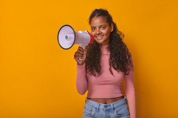 Young cute Indian woman teen with megaphone for marketing promotions in hands looks at camera dressed in casual t-shirt and jeans stands posing in orange studio. Student, schoolgirl, pupil
