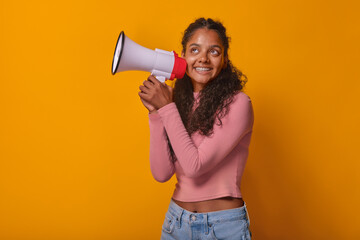 Young happy ethnic Indian woman teen with megaphone in hands looks up dreamily coming up with new slogan for advertising company or promotional event stands on plain orange background.