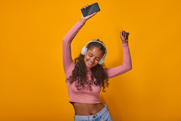 Young beautiful Indian woman dances holding mobile phone in hands and listening to music in headphones and dreams of participating in TV show with professional dancers stands in orange studio.