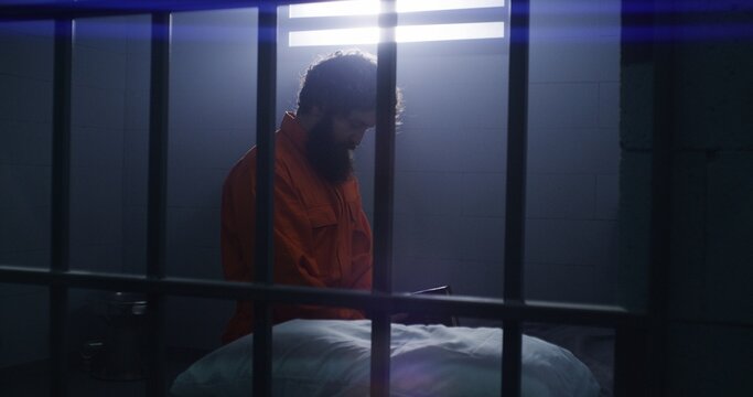Believing male prisoner in orange uniform kneels near the bed, prays to God in prison cell holding Bible. Criminal serves imprisonment term for crime in jail or detention center. Faith in God concept.