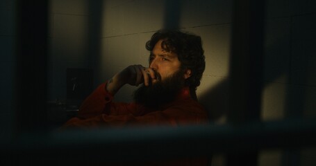 Depressed prisoner in orange uniform sits on bed, smokes cigarette in prison cell. Criminal, inmate serves imprisonment term for crime in jail. Sun shines through barred window. Correctional facility.