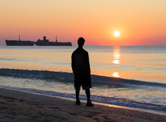 Young man admires the sunrise at the sea. Man on the seashore, the shipwreck and the sunrise.
