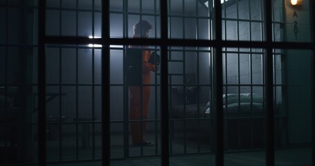 Prisoner in orange uniform kneels near bed, prays to God in prison cell holding Bible. Jailer walks, watches criminal. Inmate serves imprisonment term in jail. View through metal bars. Faith in God.