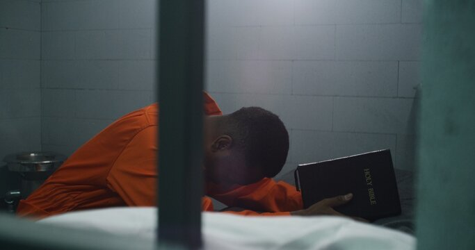 Religious African American prisoner in orange uniform kneels near the bed, prays to God in prison cell holding Bible. Male criminal serves imprisonment term in jail or detention center. Faith in God.