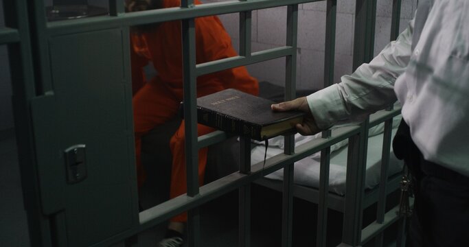 Prison officer gives Bible to male prisoner in orange uniform. Criminal sits on bed in prison cell, reads. Offender serves imprisonment term for crime in jail. Detention center. Faith in God concept.