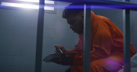 African American man sits on bed, eats dinner, serves imprisonment term for crime. Prisoner in orange uniform gives food from serving trolley to criminal in jail cell. Prison or correctional facility.