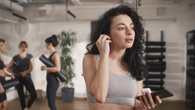 Well looking young Caucasian girl wearing earpods and searching for playlist with favorite music on her phone. Women of fitness group actively communicating before training in background.