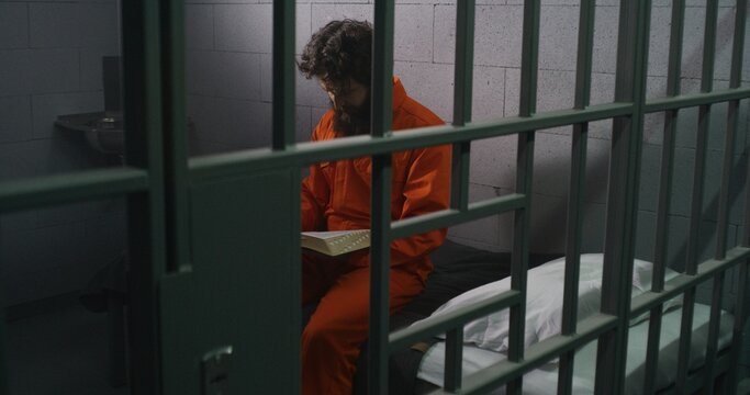 Male prisoner in orange uniform sits on bed, reads Bible, prays, looks at barred window in prison cell. Inmate serves imprisonment term for crime in jail. Correctional facility. Faith in God concept.