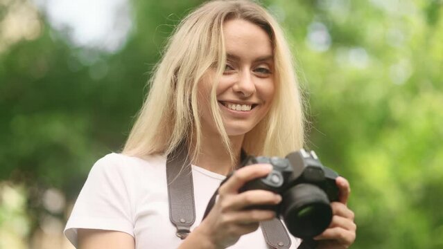 Portrait of pretty young blond woman photographer hold digital camera and looking at camera at green park Charming young tourist female taking pictures outdoors