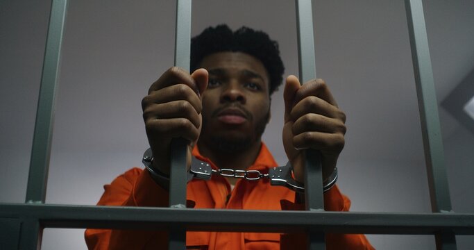 African American man in orange uniform keeps hands in handcuffs on jail cell bars. Depressed murderer serves imprisonment term in prison. Guilty prisoner in correctional facility or detention center.