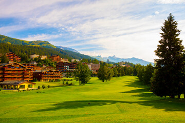 Village with House and Hotel with Panoramic View over Mountain with Clouds and Golf Course in a Sunny Summer Day in Crans Montana, Valais in Switzerland.
