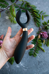 A black toy for adults in a woman's hand. The background is concrete with flowers. - 633694013