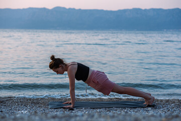A young girl in a black tank top and pink shorts doing yoga on the sea beach. Yoga at sunset overlooking the beautiful Adriatic Sea and green shores. Photo promotes a healthy lifestyle, sports, yoga.