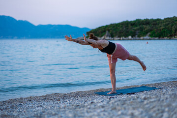 A young girl in a black tank top and pink shorts doing yoga on the sea beach. Yoga at sunset overlooking the beautiful Adriatic Sea and green shores. Photo promotes a healthy lifestyle, sports, yoga.