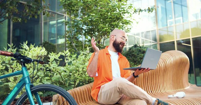 Smiling man freelancer in glasses working remotely online with headset and laptop. Young businessman talking on a video call using laptop while sitting on bench near an office building.
